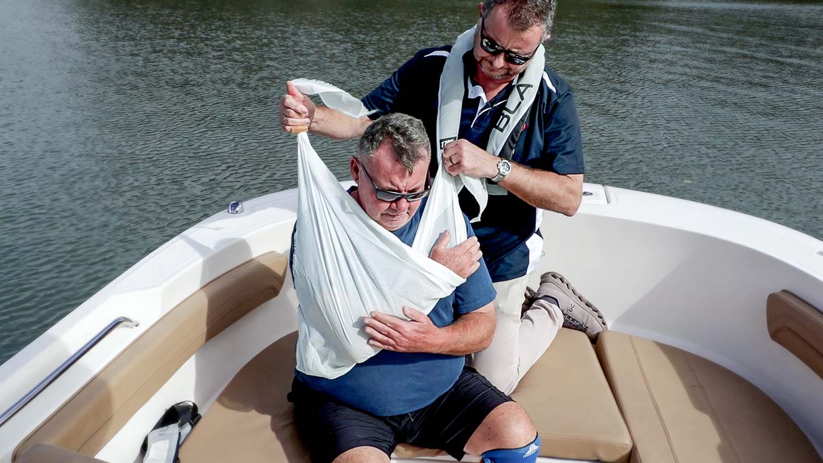 Marine First Aid responder applying sling to dislocated arm of casualty on bow of boat, with water behind